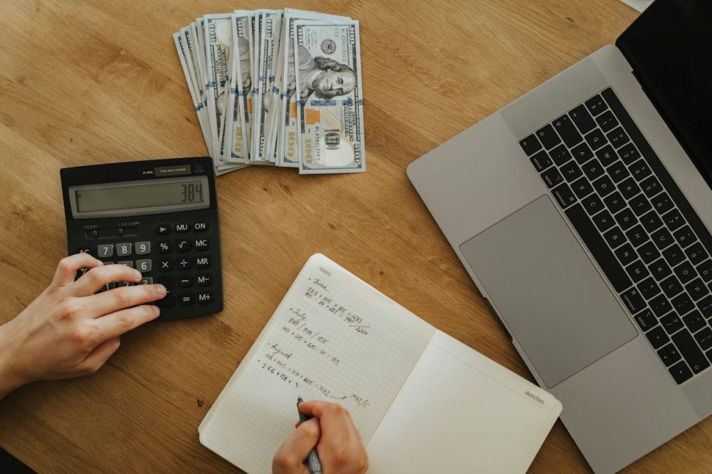 Hands using calculator and notebook, planning finances, with cash and laptop on wooden table.